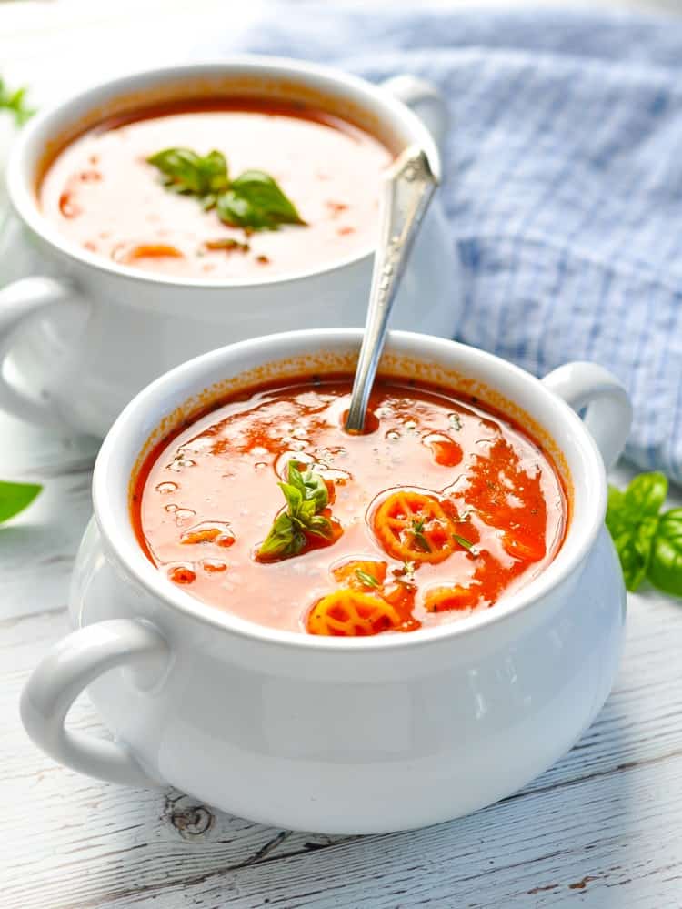 A white bowl filled with marinara soup and topped with pasta wheels and basil