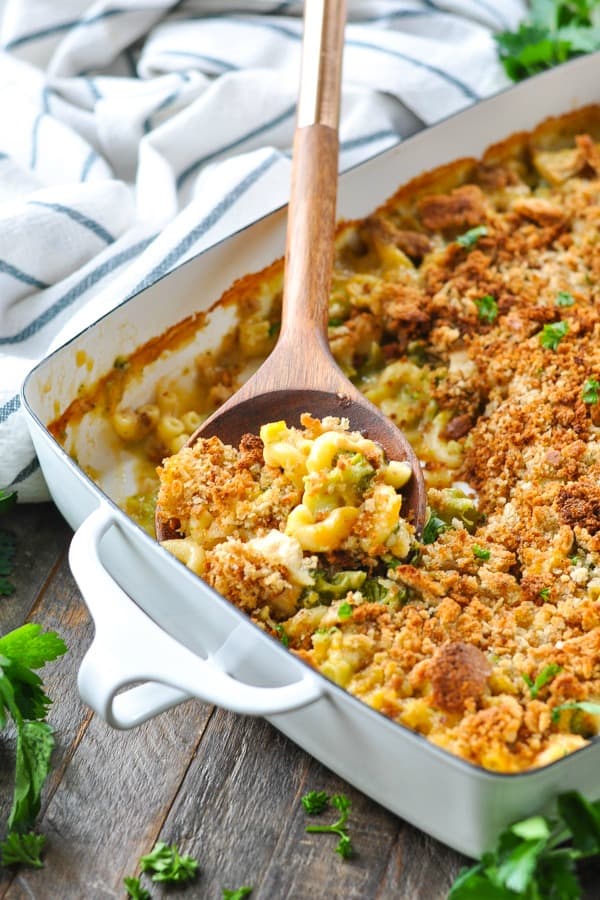A wooden spoon scoops a spoonful of cheesy chicken and broccoli casserole from a white ceramic casserole dish.
