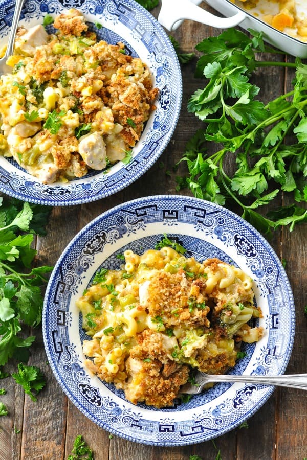 Two bowls filled with cheesy broccoli and chicken casserole, topped with stuffing. Bunches of fresh parsley surround the bowls.