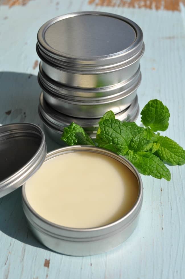 Homemade lip balm in small round metal tins. One tin has the lid removed, revealing the light yellow colored lip balm. Fresh mint leaves sit beside the tins.