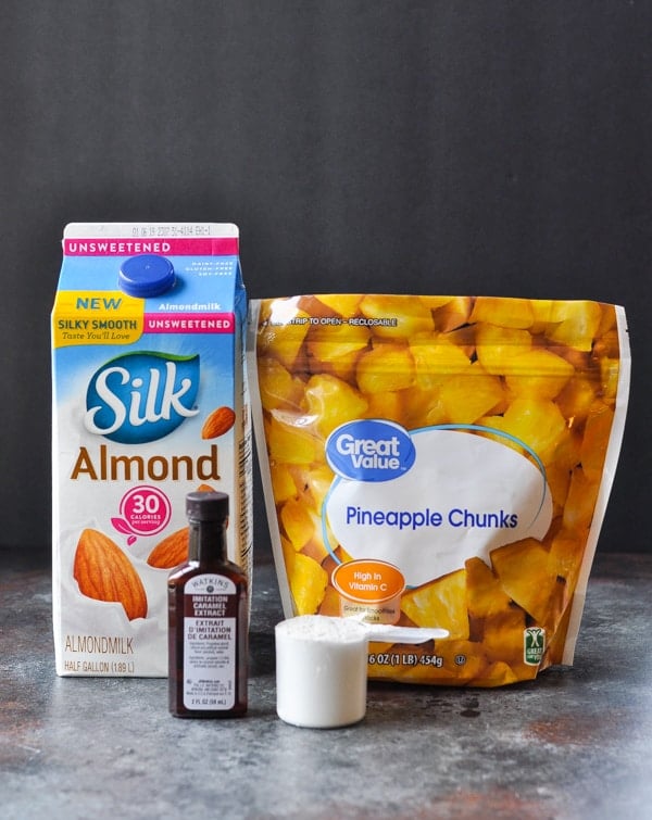 The ingredients needed to make a healthy pineapple protein shake -- almond milk, caramel flavoring, frozen pineapple chunks, and a scoop of protein powder.