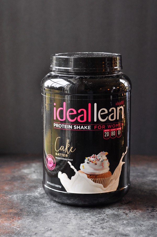 A large tub of IdealLean cake batter flavored protein powder. The container is all black with pink and white lettering, and a picture of a cupcake splashing into creamy milk.