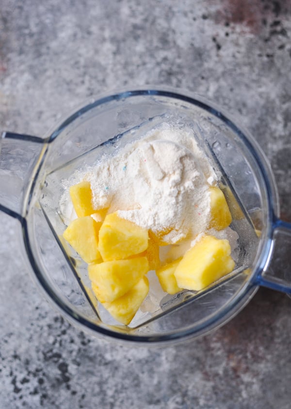 An overhead image of pineapple chunks, protein powder, milk, and ice in a blender.