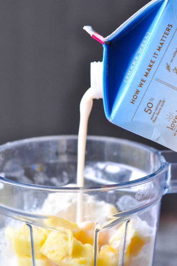 A person pours almond milk from a blue carton into a blender containing the ingredients for a healthy pineapple protein smoothie.