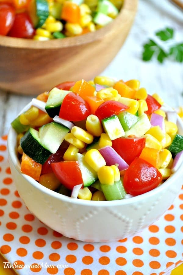 A small white bowl filled with a healthy veggie salad, made up of chopped cucumbers, peppers, onions, tomatoes, and corn.