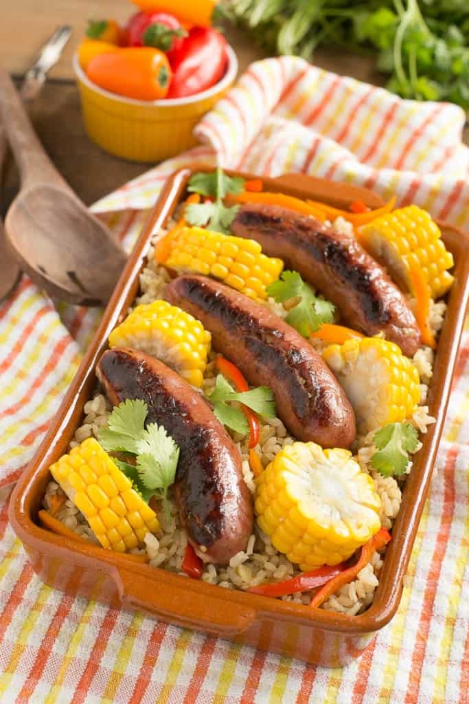 A one-pot sausage and rice bake served with corn on the cob served in a brown casserole dish.