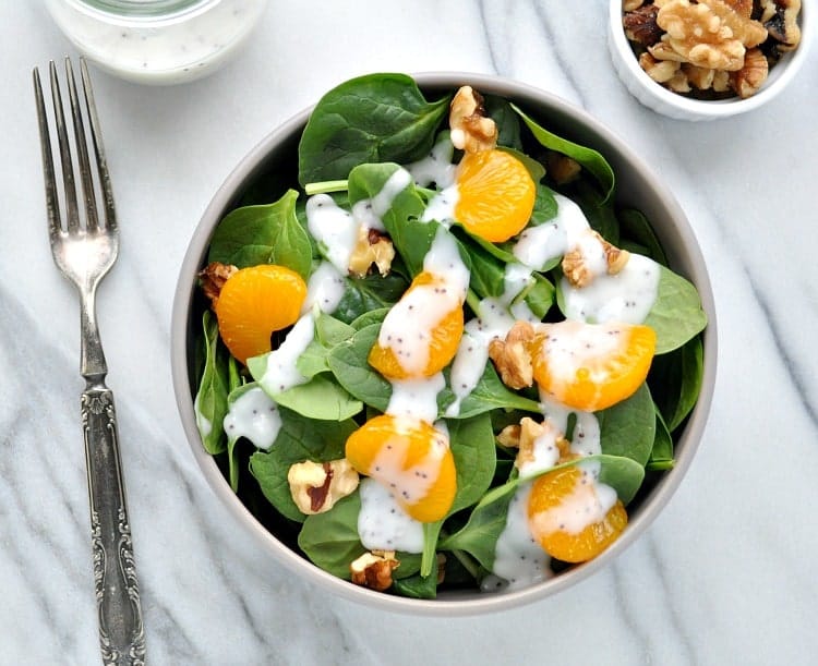An overhead shot of a bowl of salad - spinach leaves topped with mandarin slices, walnuts, and poppyseed dressing, - on a white marble countertop served with a fork and a small bowl of walnuts.