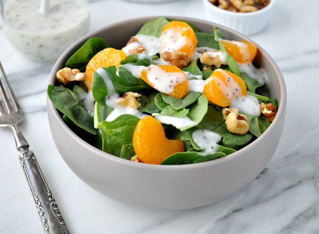 A white bowl filled with a spinach salad topped with mandarin orange slices, walnuts, and a creamy poppyseed dressing.
