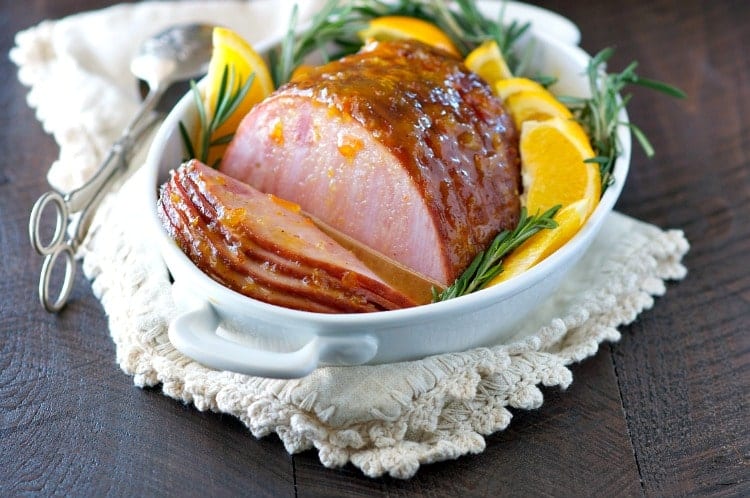 A glazed baked ham in a white dish with wedges or orange and rosemary