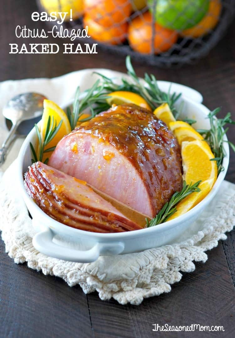 A glazed baked ham in a white dish garnished with orange wedges and rosemary