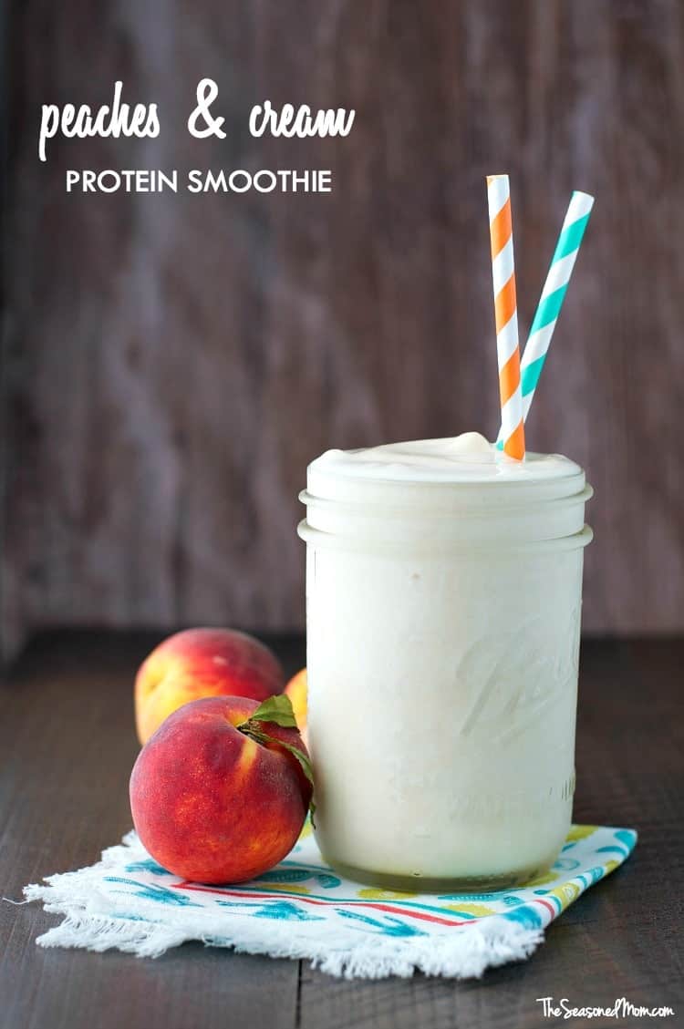 A peaches and cream protein smoothie in a glass with two straws and peaches at the side