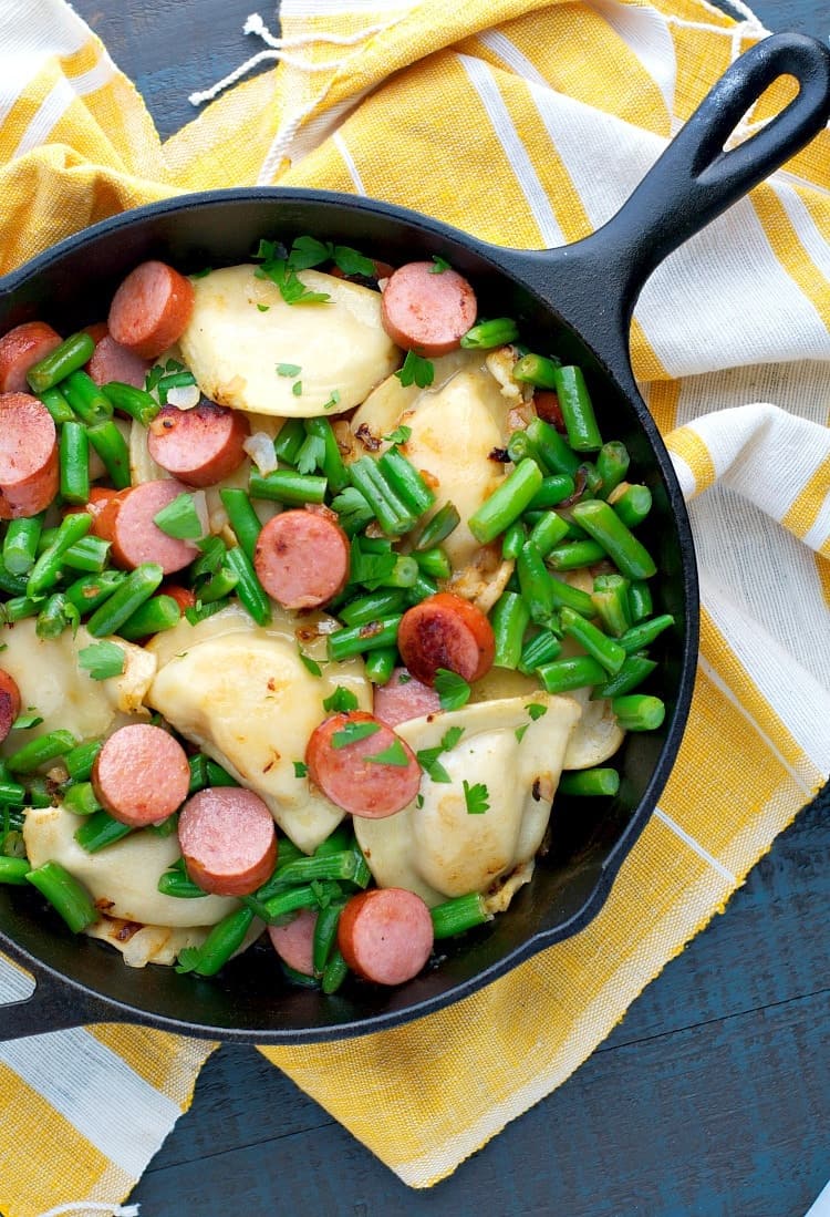 An overhead image of a kielbasa and pierogies skillet with onions and green beans, served over a yellow and white tea towel.