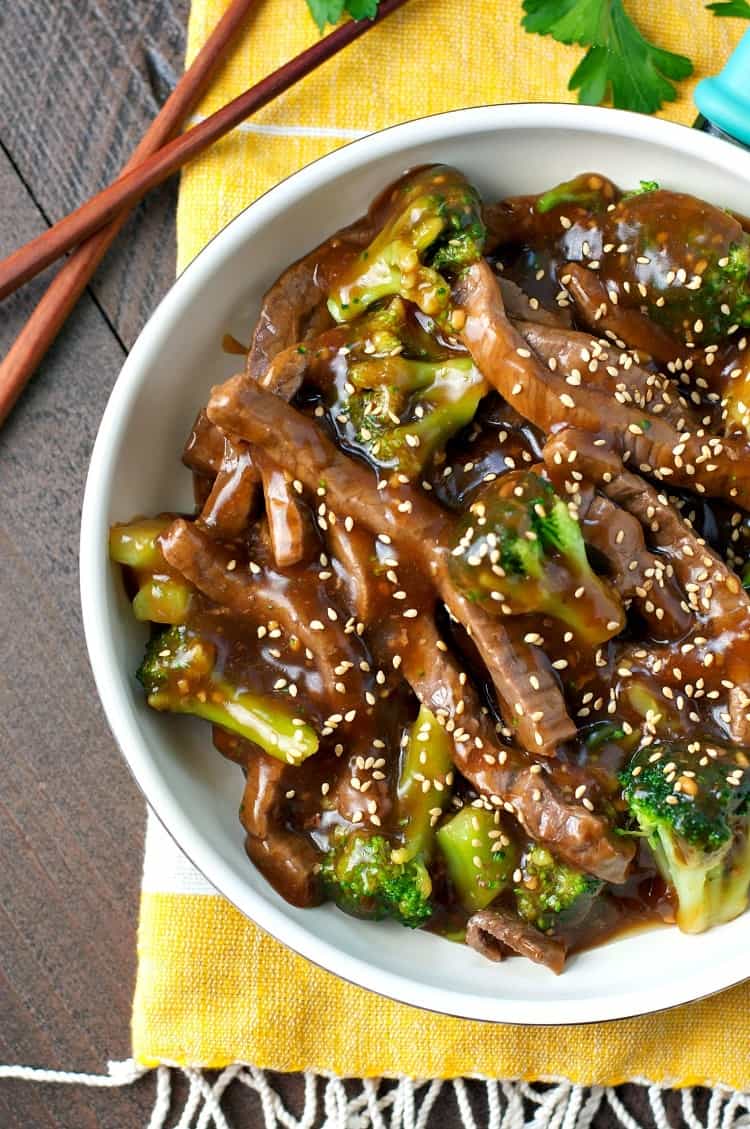 An overhead image of a large bowl of Mongolian beef and broccoli, served on a table with a white and yellow striped tea towel and a set of bamboo chopsticks.