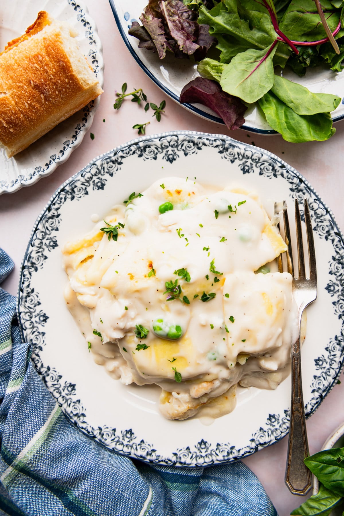 Overhead shot of chicken ravioli alfredo on a blue and white plate with a side salad and bread.