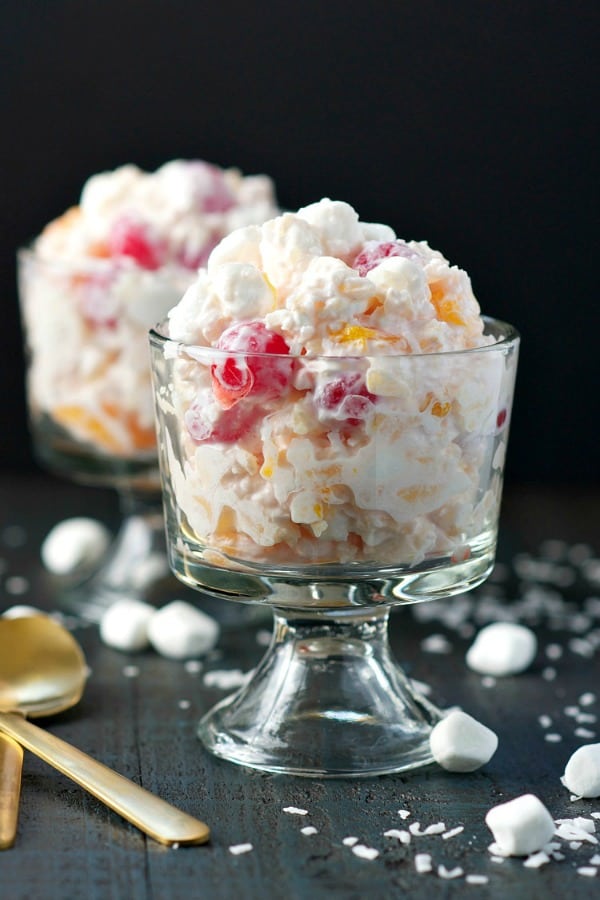 Two bowls of ambrosia salad on a dark wood surface surrounded by coconut and marshmallows