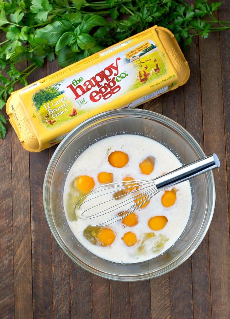 An overhead image of eggs mixed in a glass bowl with whole milk. A yellow carton of eggs sits on the table next to the mixing bowl.