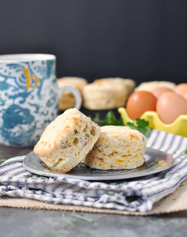 Two cheddar biscuits on a plate with a mug and eggs in the background