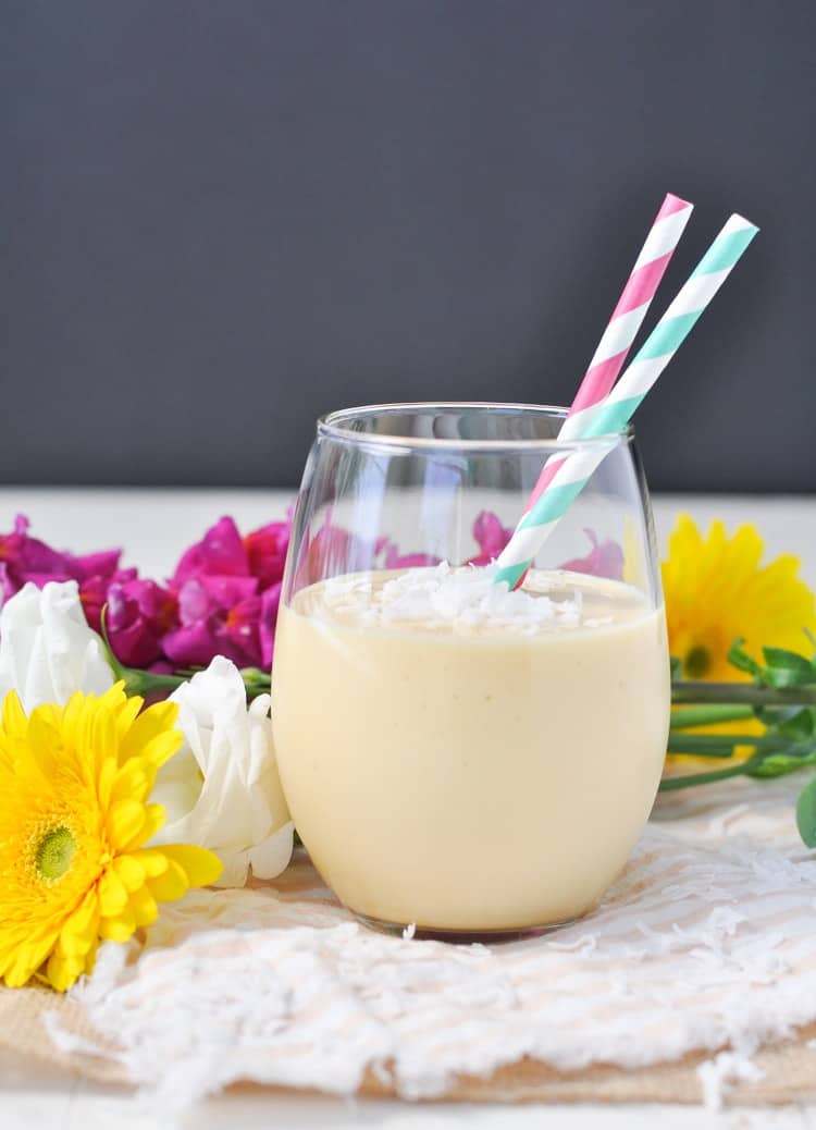 A glass filled with a coconut smoothie with two straws and flowers in the background