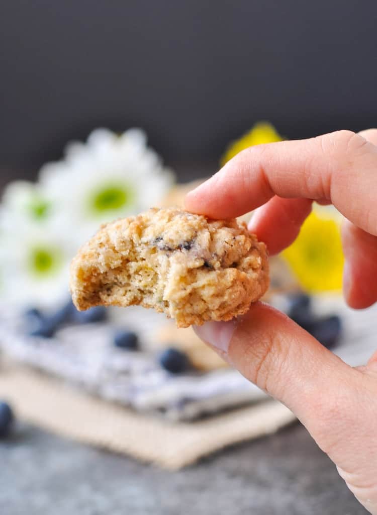 A person holds a single blueberry oatmeal muffin cookie between two fingers. The cookie has a small bite taken out of it.