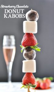 A close up of a strawberry chocolate donut kabob with a glass of wine in the background