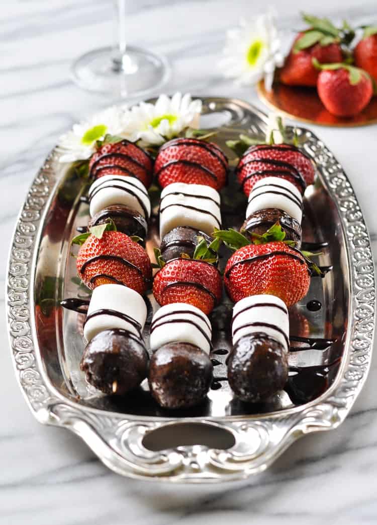 A photo of three strawberry chocolate donut kabobs on a silver plate with a bowl of strawberries in the background
