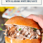Hand holding slow cooker pulled pork sandwich with alabama white barbecue sauce and text title box at top