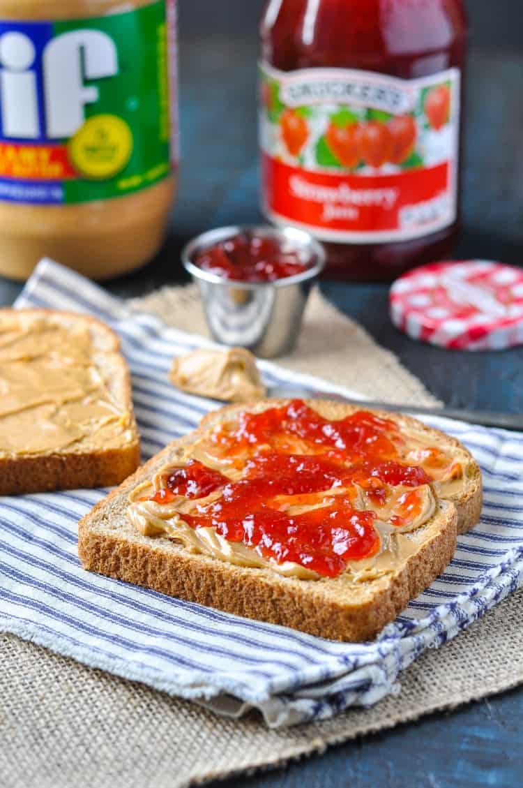 An open peanut butter and jelly sandwich on a striped dish cloth with condiments in the background