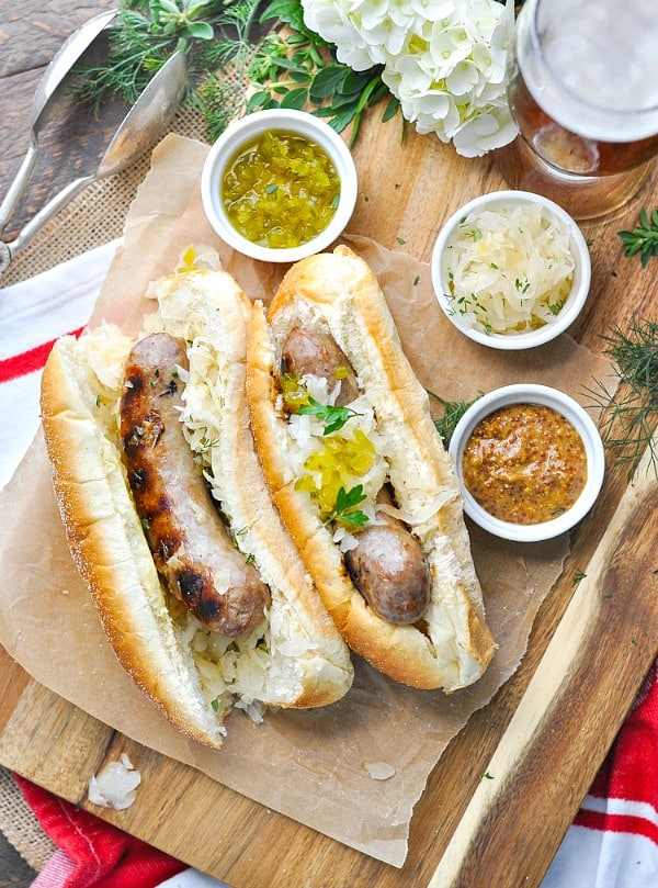 An overhead shot of beer brats on a wooden board with sauces and onions