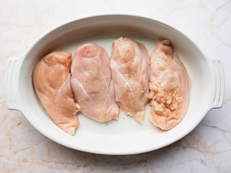 Chicken breasts arranged in a white oval baking dish and seasoned with salt and pepper.