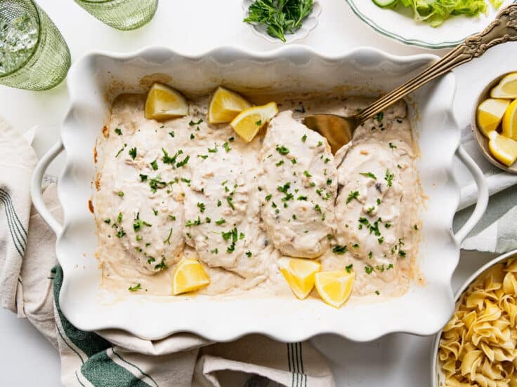 Horizontal overhead shot of a baked dish full of cream of mushroom chicken garnished with parsley and lemon wedges.