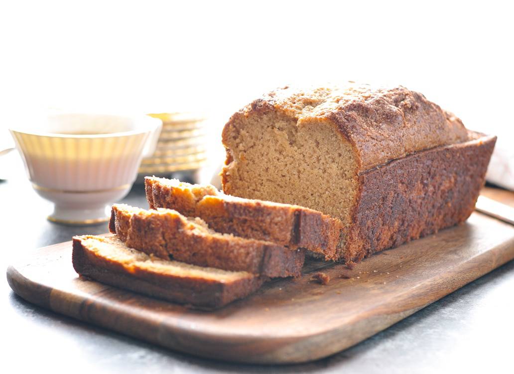 A photo of a loaf of amish friendship bread cut into slices on a chopping board with cups of coffee in the background
