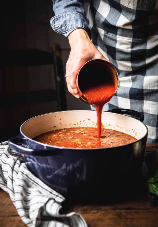 A woman pours a big can of tomato sauce into a dutch oven, filled with spaghetti sauce.