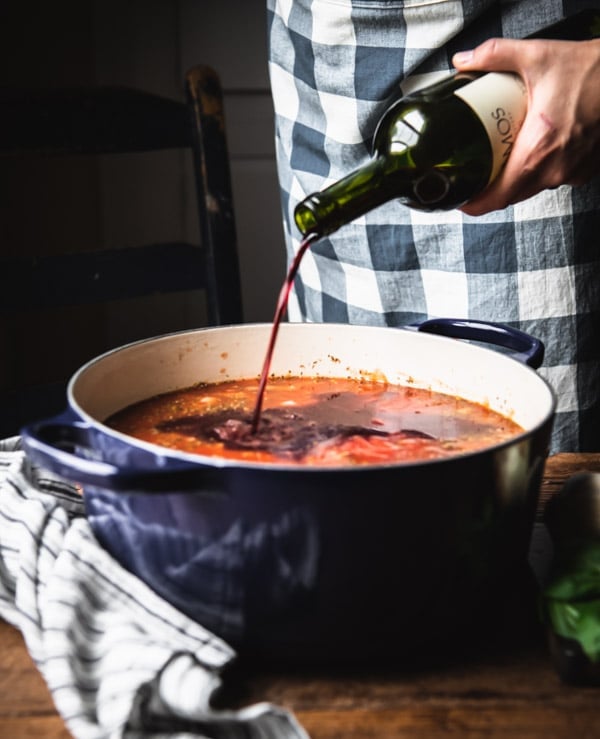 A woman pours red wine from a bottle into a blue Dutch oven filled with cooking spaghetti sauce.