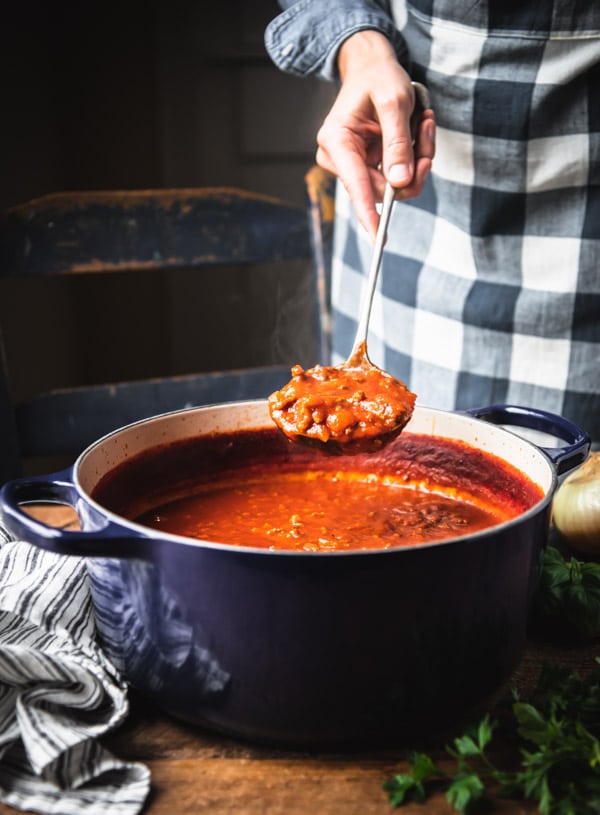 A woman holds a ladleful of homemade stovetop spaghetti sauce, scooped from a large blue Dutch oven.