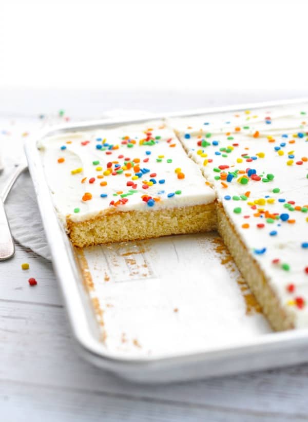 Two slices are removed from a pan of white Texas sheet cake. The vanilla sheet cake is topped with a thin layer of vanilla buttercream and rainbow confetti sprinkles.