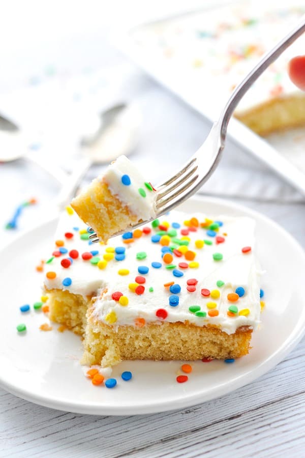 A silver fork lifts a small bite of white Texas sheet cake from a plate. The slice of vanilla cake is topped with vanilla frosting and colorful sprinkles.