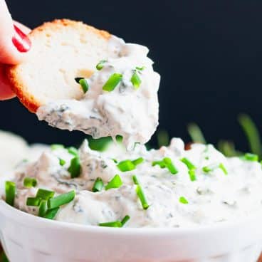 Square close up shot of a hand dipping a bagel chip into a bowl of 4 ingredient spinach dip.