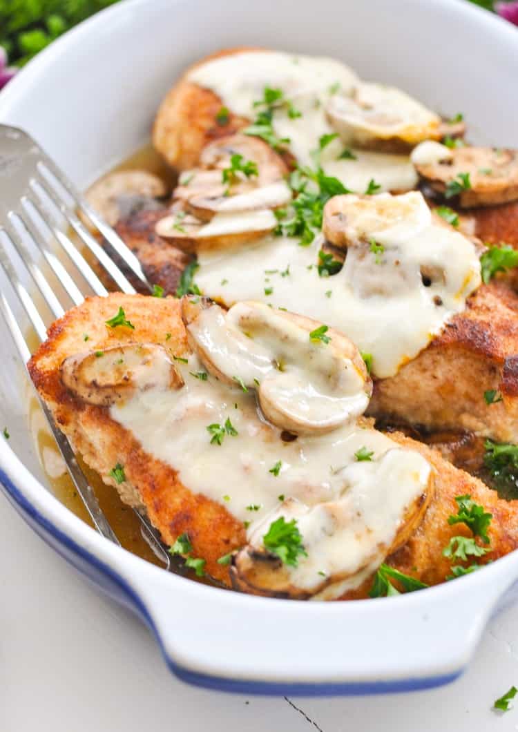 Close up photo of golden baked chicken breasts topped with mushrooms and melted cheese in a casserole dish.