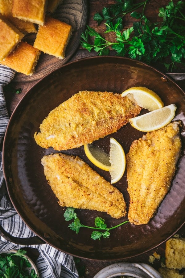 Close overhead shot of crispy fried catfish fillets in a cast iron skillet with lemon wedges and parsley