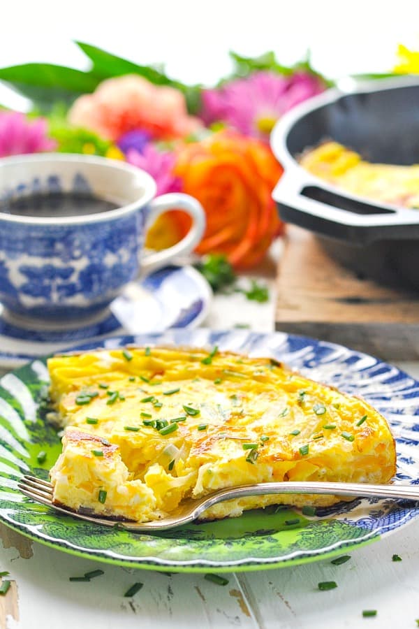 A photo of a slice of easy frittata made with cheddar cheese and chives on a blue plate with a cup of coffee in the background