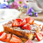 Slice of Easy French Toast Casserole on a white plate topped with fresh strawberries and pour of maple syrup