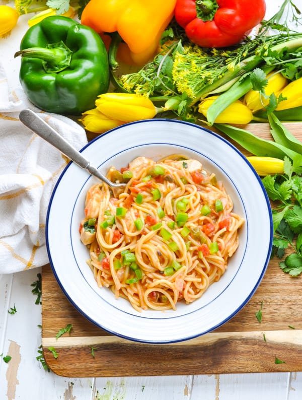 An overhead image of a bowl of baked chicken pasta, served on a cutting board surrounded by fresh bell peppers and bunches of yellow flowers.