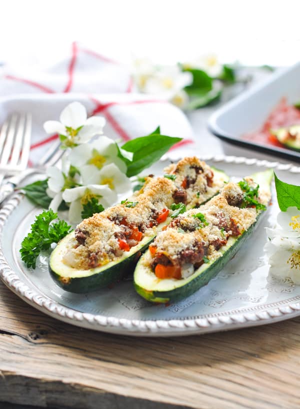 Two baked stuffed zucchini boats filled with a hearty meat and vegetable filling sit on a white plate surrounded by blooms of white flowers.