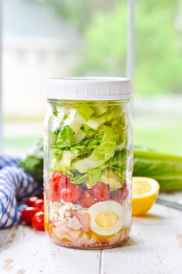 A side shot of a Mason Jar Turkey Cobb Salad sitting on a white surface in front of a window