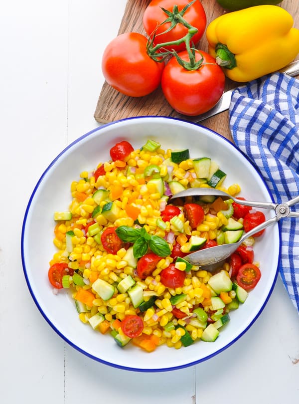 An overhead image of a large bowl of fresh corn salad with tomatoes, zucchini, peppers, and onions. The dish is served with silver salad tongs, alongside on the vine tomatoes.