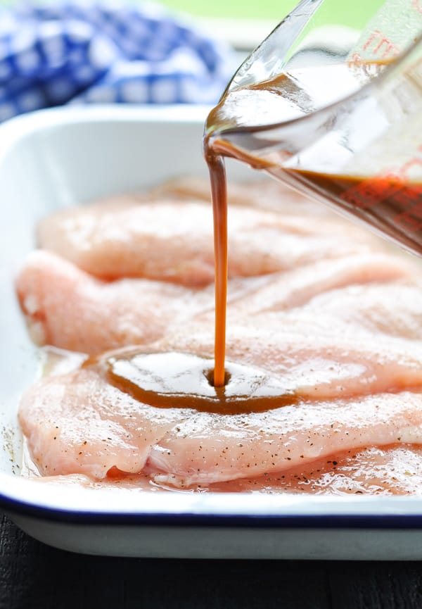 Pouring balsamic sauce from a glass measuring cup over raw, seasoned chicken breast in a baking dish.
