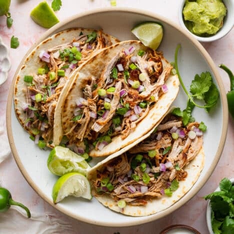 Square overhead shot of carnitas in tacos on a white plate with fresh cilantro, limes, and onions for garnish.