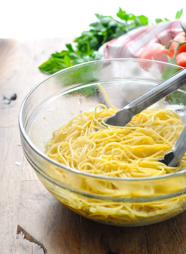 A glass bowl filled with freshly cooked spaghetti pasta, coated in a garlic-infused olive oil and tossed with metal tongs.