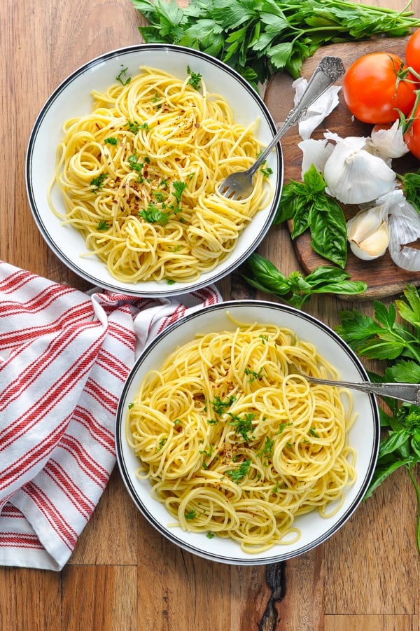 Two full bowls of spaghetti aglio e olio served with silver forks, surrounded by fresh ingredients - basil leaves, cloves of garlic, and vine-ripe tomatoes.