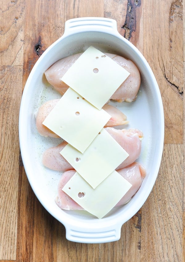 Overhead image of raw boneless skinless chicken breasts in a white dish topped with slices of Swiss cheese